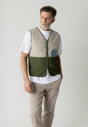 Mand med gråt skæg iført beige og grøn vest med lynlås over hvid T-shirt og lyse bukser, stående mod en ensfarvet lys baggrund.