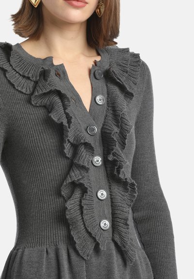 Cardigan grigio a maglia con bordo arricciato sul davanti, bottoni tondi con texture dettagliate e vita aderente che si allarga verso il fondo.
