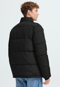 Sort pufferjakke med quiltet design, høj krave og løs pasform. Stoffet ser glat ud med elastiske manchetter, parret med blå jeans.