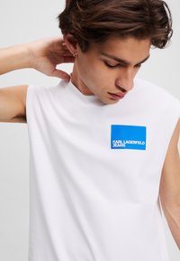 Tricou alb fără mâneci cu un patch pătrat albastru pe care scrie "KARL LAGERFELD JEANS" cu text alb. Textură netedă și design ajustat.