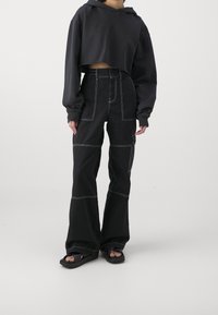 Persoon in een zwart cropped hoodie en hoge zwarte wijde broek met contrasterende witte stiksels en zwarte sandalen op een witte achtergrond.