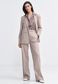 Beżowy garnitur w prążki z dwurzędową marynarką i szerokimi spodniami. Pasująca satynowa koszula ma delikatny połysk, tworząc spójną całość.