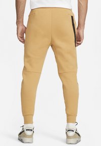 Slim-fit, mustard sweatpants with side zip pockets, smooth texture, tapered cuffs, and reinforced knee panels. Worn with athletic sneakers.