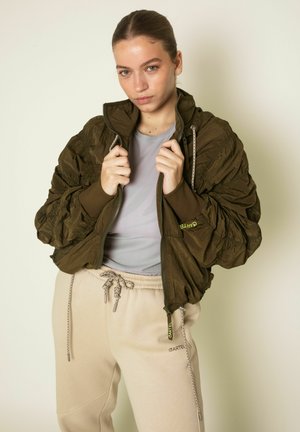 Junge Frau trägt eine olivgrüne gesteppte Jacke über einem hellgrauen Oberteil und beigen Jogginghosen, hält den Jackenkragen mit beiden Händen fest.