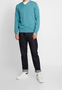 Man som bär en teal V-ringad tröja, mörka uppvinklade jeans, vita strumpor och vita sneakers med marinblå accent mot en plain bakgrund.