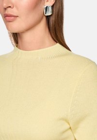 Ribbed, high-neck sweater in soft yellow fabric; paired with oversized, silver-tone square earrings with a polished finish.
