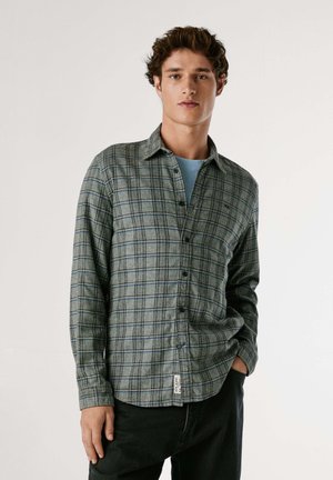 Camicia a quadri verde in tessuto morbido con chiusura a bottoni, maniche lunghe e un'etichetta sull'orlo. Indossata sopra una t-shirt azzurro chiaro.
