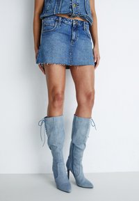 Jupe en denim mini avec un ourlet effiloché, assortie �à des bottes bleu clair jusqu'aux genoux avec des bouts pointus et des lacets réglables sur les côtés.