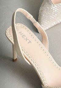 Crèmekleurige slingback pumps met een geschulpte rand op de binnenzool en een dunne stilettohak. De binnenkant is beige met een gedrukt merkje.