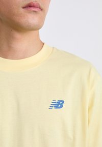 Šviesiai geltona medvilninė marškinė su apvaliu iškirpimu, ant kairiojo krūtinės puikuojasi mėlynas "NB" logotipas. Sklandi tekstūra, laisvas siluetas.