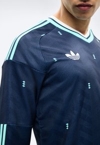 Muškarac obučen u tamnoplavu Adidas sportsku majicu sa svijetloplavim prugama i logom, krupni kadar na ramenima i prsima.