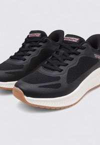 Chaussures de sport noires avec tissu en maille, superpositions texturées et une semelle intermédiaire blanche. Elles possèdent des lacets noirs et une semelle extérieure en caoutchouc marron.