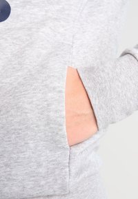 Material de sudadera gris con un bolsillo lateral, que presenta una textura sutil y un ajuste cómodo. La mano está metida en el bolsillo.