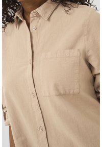 Chemise beige à boutons, en tissu doux et texturé, dotée d'une poche poitrine unique et d'un col classique, avec des manches retroussées.