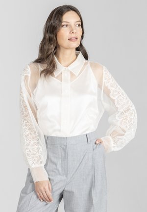 Kobieta o falujących brązowych włosach, ubrana w przezroczystą białą bluzkę z koronkowymi rękawami oraz jasnoszare spodnie z wysokim stanem, patrząca na bok.