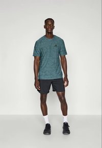 Camiseta deportiva azul verdoso de Adidas con un patrón de rayas texturizadas, con un logo negro, combinada con pantalones cortos negros y zapatillas oscuras.