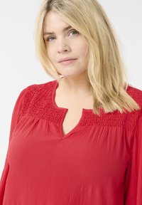 Červená blůza se smockovaným výstřihem, véčkovým výstřihem a dlouhými rukávy. Tkanina má texturovaný vzhled, který doplňuje její uvolněný střih.