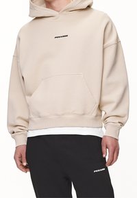 Beżowa bluza z kapturem z przednią kieszenią, ściągaczami przy rękawach i u dołu, z małym czarnym logo "PEGADOR" na klatce piersiowej. Nosi ją na białej koszuli.