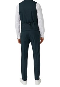 Man in een donkerblauwe pantalon en bijpassend vest over een wit overhemd met witte sneakers, gezien vanaf de achterkant.
