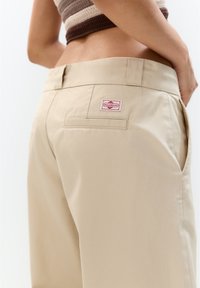 Beige bukser lavet af glat stof med lige snit, som har en baglomme og et rødt "FULL&BEAR" mærkat.