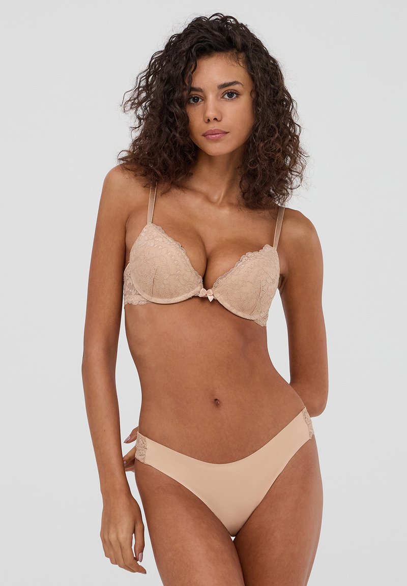 Reggiseno color nudo con pizzo, coppe imbottite e spalline regolabili, abbinato a slip in stile bikini con dettagli in pizzo.