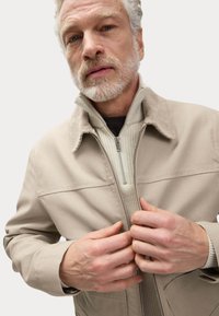 Giacca zip-up color marrone chiaro con colletto in velluto a coste, indossata sopra un maglione a coste di un beige leggero. Il modello stringe la zip della giacca.