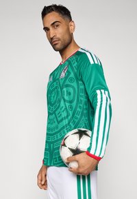 Πράσινη ποδοσφαιρική μπλούζα μακρυμάνικη με γεωμετρικό σχέδιο, με κόκκινες και λευκές λεπτομέρειες, και λογότυπο στο στήθος. Κρατάει μια μπάλα ποδοσφαίρου.