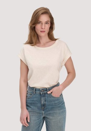 Junge Frau mit hellbraunen Haaren, die ein cremefarbenes Kurzarmshirt trägt, das in eine hoch taillierte blaue Jeans gesteckt ist, stehend vor einem einfarbigen Hintergrund.