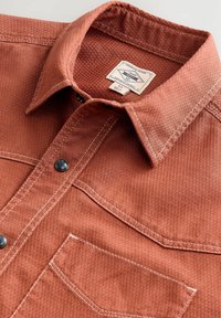 Chemise couleur rouille avec tissu texturé, col pointu, bouton snap noir et couture blanche contrastante, dotée d'une poche poitrine et d'une étiquette.