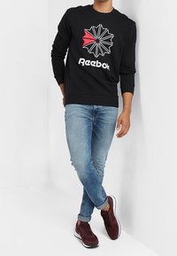 Svart sweatshirt med stjärndesign i vitt och rött, kombinerad med ljusblå jeans och vinröda sneakers, stående mot en vit vägg.