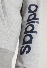 Sweatshirt gris avec une texture de tissu lisse, présentant un grand logo "adidas" bleu marine le long de la manche et un design de capuche standard.