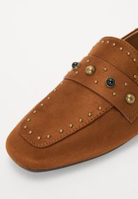 Mocassins en cuir suédé marron ornés de clous métalliques dorés et noirs, dotés d'une semelle plate et d'un design à bout arrondi. Les accents incluent une sangle décorative.