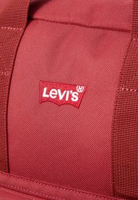Červená tkanina s textúrovaným povrchom, na ktorej je vyšívané biele logo "Levi's" v kontrastnej záplate. Dizajn zahŕňa hrubé švy a pevné popruhy.