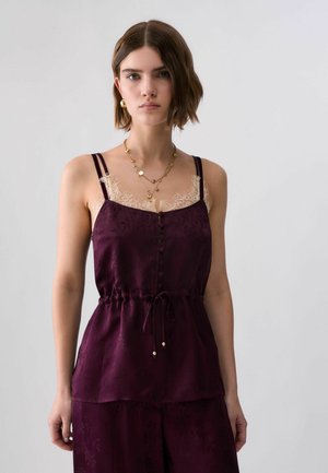 Joven con pelo corto y castaño que lleva una camiseta sin mangas de color púrpura oscuro con ribete de encaje, joyas doradas y pantalones a juego sobre un fondo liso.