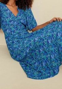 Robe maxi bleue à fleurs avec un décolleté en V, des manches courtes et un motif tourbillonnant de nuances de vert, de bleu et d'accents blancs. Texture de tissu douce et fluide.