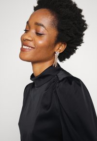 Blusa preta de gola alta com mangas bufantes, textura suave e ombros estruturados. Modelo usa brincos pendentes com detalhes transparentes.