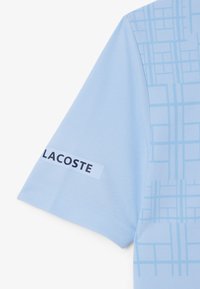 Polo bleu clair avec un motif de grille texturé. Comprend une étiquette blanche avec "LACOSTE" imprimé en lettres noires grasses sur la manche.