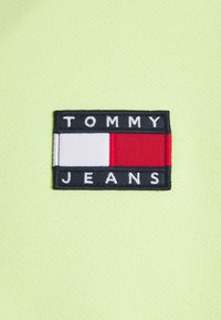 Parche rectangular negro con secciones blanca, roja y azul, que presenta el texto "TOMMY JEANS" en blanco sobre un fondo de tela amarillo brillante.