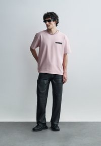 Uomo che indossa una T-shirt oversize rosa chiaro, pantaloni in pelle neri, scarpe da ginnastica nere, occhiali da sole scuri, in piedi contro un muro grigio chiaro uniforme.