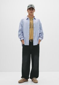 Lysblå stribet skjorte med knapper over en sennepsgul grafisk t-shirt, parret med løse sorte jeans og brune sneakers; grå kasket.