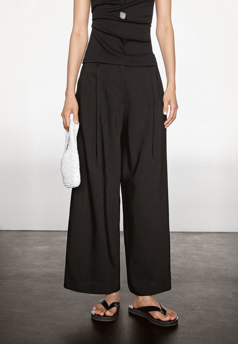 Zwarte wijduitlopende broek van lichtgewicht stof, gecombineerd met een strakke zwarte top. Een witte geweven tas en zwarte slippers maken de look compleet.
