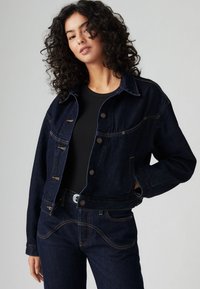 Tamno traper jakna s kraćim krojem, dva džepa na prsima i bakrenim šavovima. Kombinirana s crnim rebrastim topom i odgovarajućim trapericama.