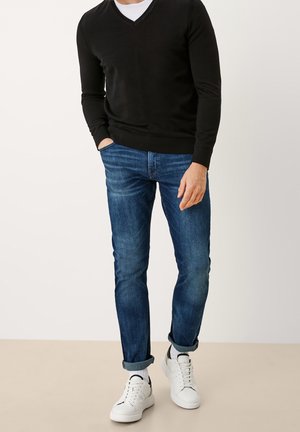 Uomo con maglione nero a scollo a V, jeans blu risvoltati, calzini bianchi e scarpe da ginnastica bianche in piedi su uno sfondo chiaro.