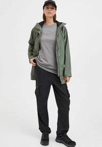 Green waterproof jacket over a gray shirt, paired with black cargo pants and black shoes. The jacket has a hood and front zip.
