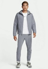 Bărbat în picioare, îmbrăcat într-o jachetă cu glugă gri, pantaloni gri asortați, un tricou alb Nike și încălțări atletice gri și alb pe un fundal alb.