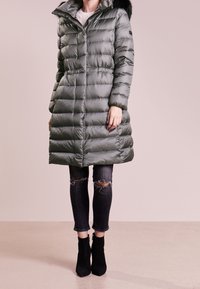 Nafoukaná zelená parka s vysokým límcem, odnímatelným kožíškem a pasem se zavazováním, doplněná o potrhané černé džíny a kotníkové boty.