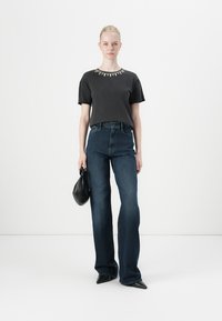 Cămașă neagră cu mâneci scurte, cu decolteu embellisat, asortată cu blugi evazați cu talie înaltă de culoare albastru închis. Modelul ține o geantă mică neagră.