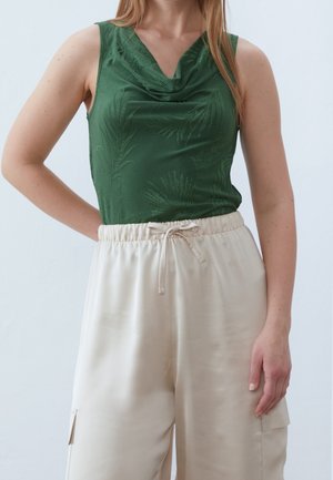 Femme portant un haut vert sans manches à motif de feuilles et un pantalon beige taille haute avec cordon et poches latérales sur un fond clair uni.