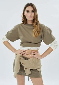 Beżowy krótki sweter z krótkimi rękawami, nałożony na biały długi t-shirt, w połączeniu z dopasowaną beżową mini spódnicą i związanym swetrem.