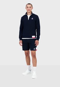 Felpa blu con zip e colletto bianco, abbinata a pantaloni corti blu con accenti rossi e bianchi. Indossata con scarpe da ginnastica bianche e calze.